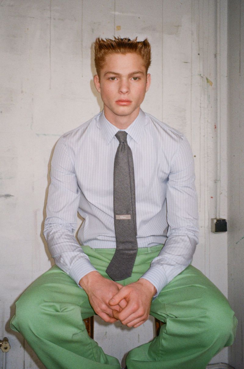 Jasper Hodges seated wearing a light pinstriped dress shirt, grey necktie, and green trousers against a studio wall.