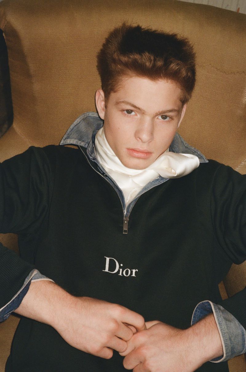 Jasper Hodges wearing a black Dior quarter-zip pullover with a white neck scarf while reclining in an armchair.
