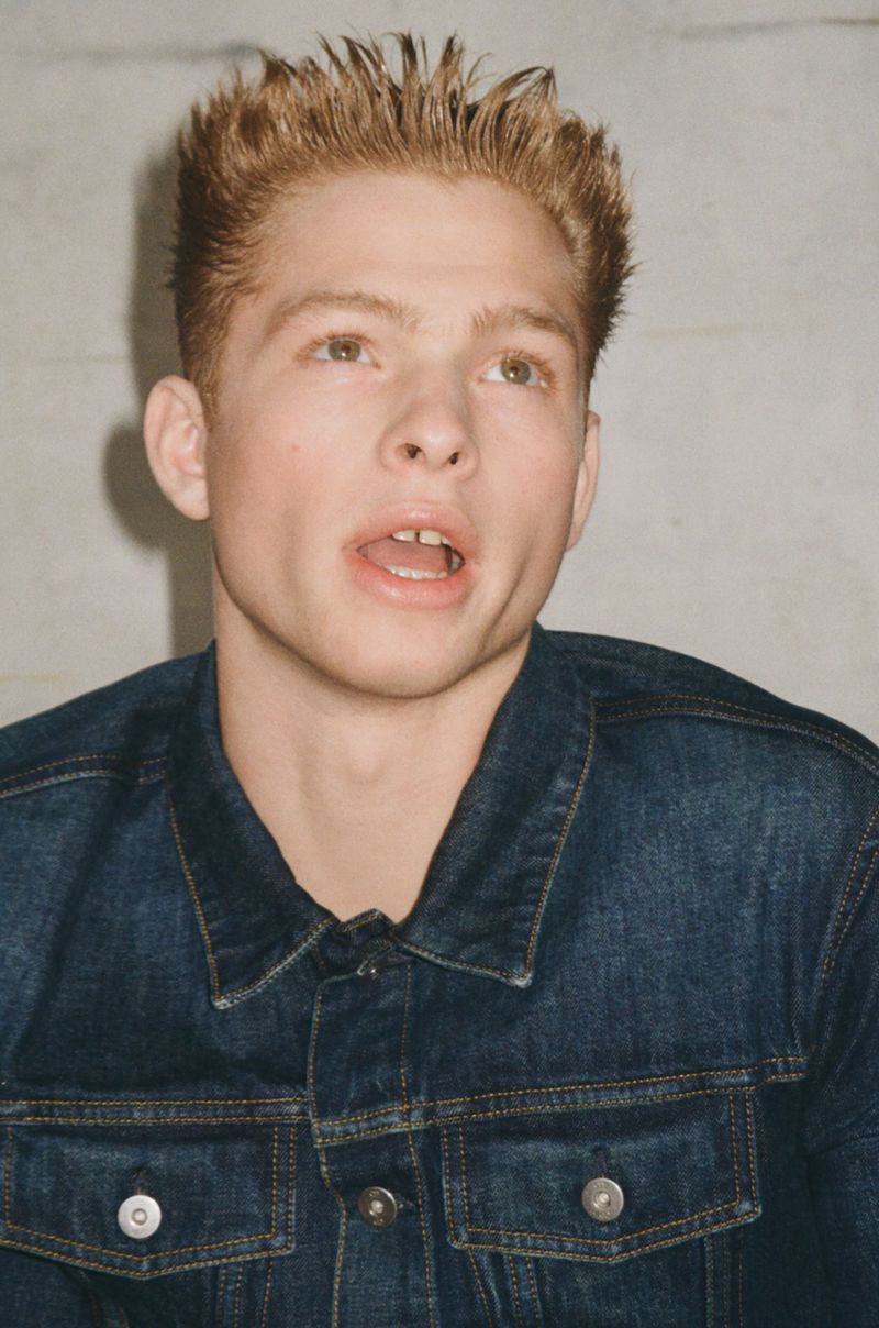Model Jasper Hodges wearing a dark blue denim jacket looking upward against a light neutral background.