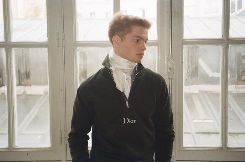 Model Jasper Hodges in a black Dior quarter-zip sweater with a white neck scarf in front of tall multi-pane windows.