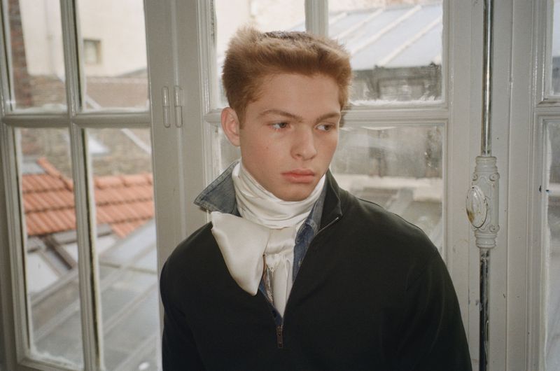 Young male model with reddish-brown hair wearing a dark jacket and white satin neck scarf standing by a multi-pane window.
