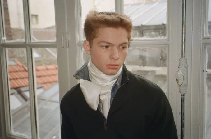 Young male model with reddish-brown hair wearing a dark jacket and white satin neck scarf standing by a multi-pane window.