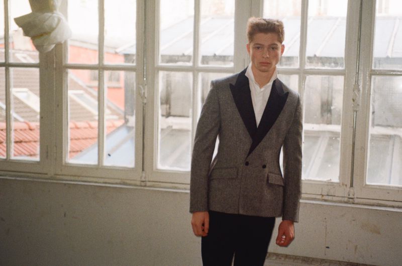 Model Jasper Hodges standing indoors in a grey double-breasted blazer with black lapels in front of tall multi-pane windows.