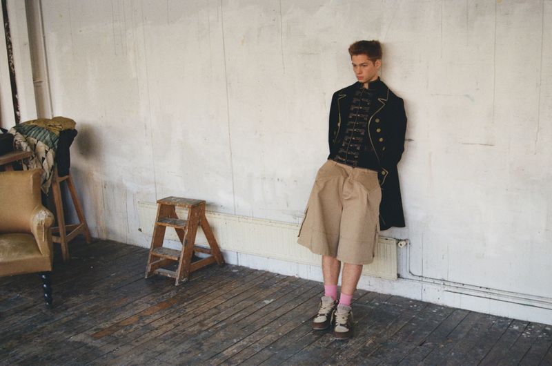 Model Jasper Hodges wearing an ornate black military-style jacket and wide beige shorts leaning against an off-white wall.
