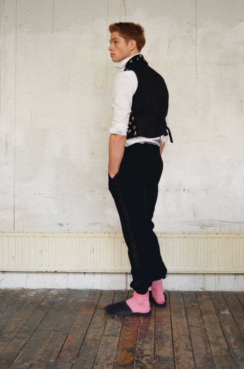 Model Jasper Hodges standing in profile wearing a black vest, white collared shirt, black trousers, and pink socks on wooden floors.