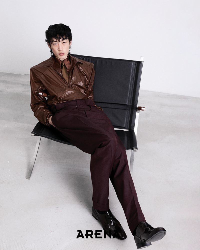 Model seated in a black leather chair wearing a brown glossy jacket and burgundy trousers against a white backdrop.