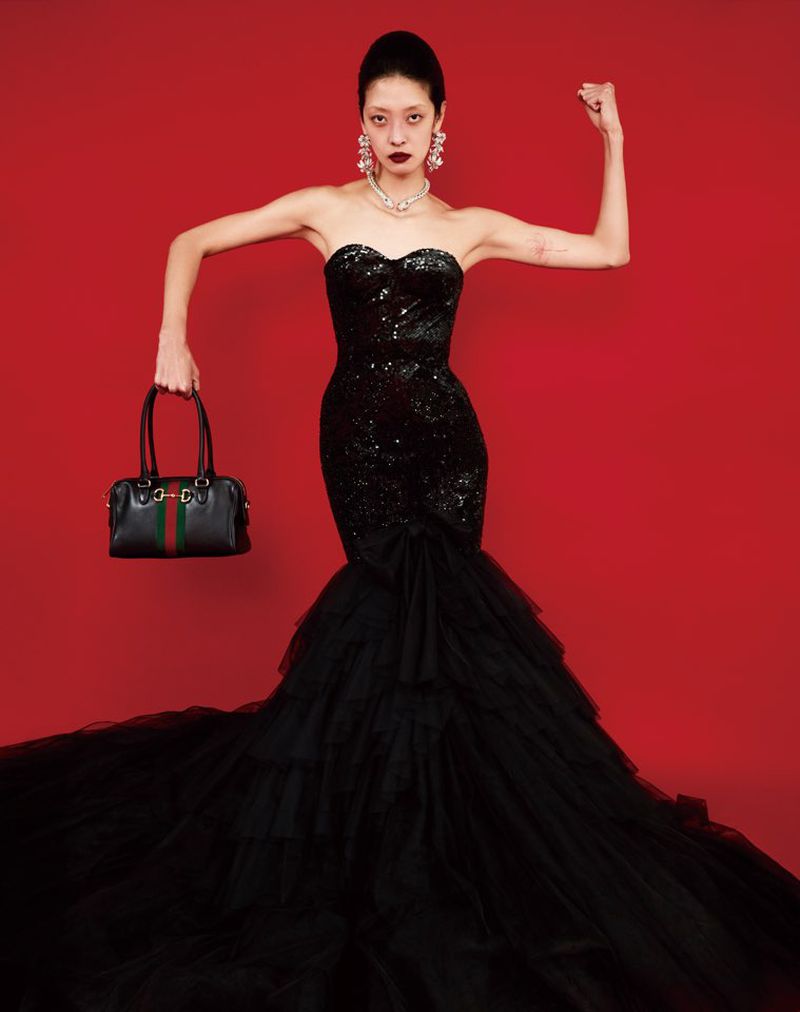Model So Young Kang posing in a black strapless mermaid gown while holding a handbag against a red background.