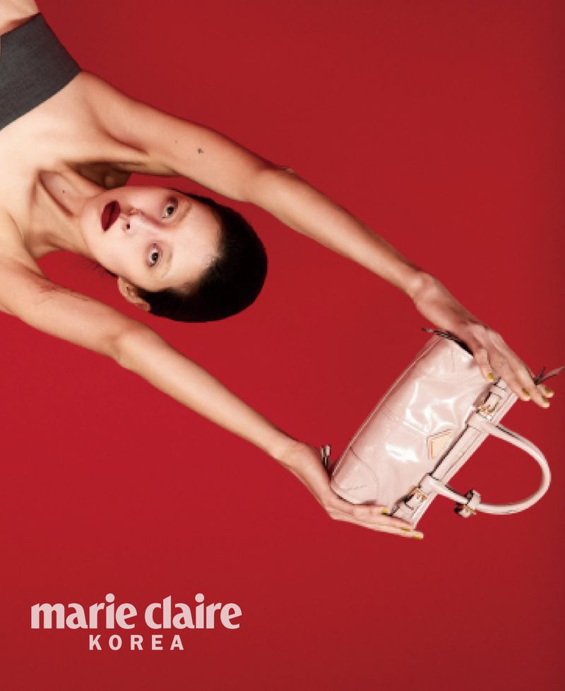 Model So Young Kang posed inverted holding a glossy pale pink handbag against a solid vibrant red background for Marie Claire Korea.