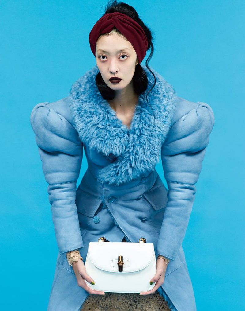 Model So Young Kang posing in an exaggerated blue coat and red headband holding a white handbag against a blue backdrop.