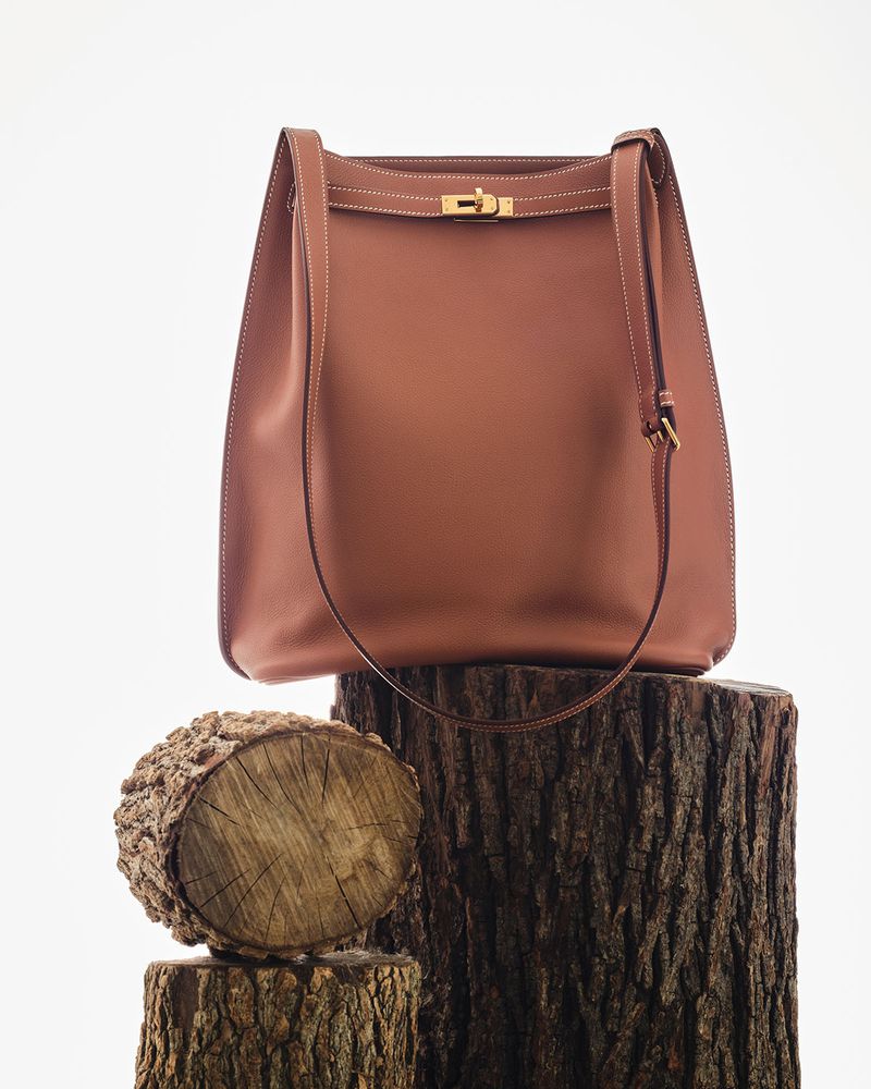 Brown leather handbag with gold clasp resting on stacked natural tree logs against a plain white background.