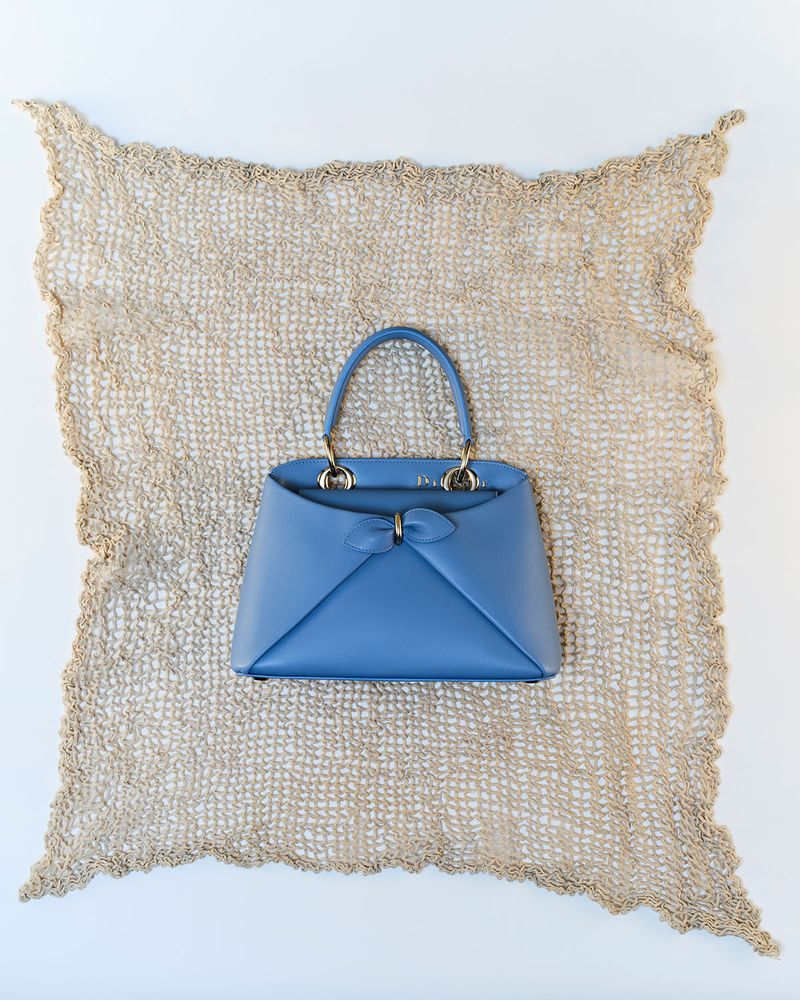 Cornflower blue leather top handle handbag displayed on a beige crochet mat against a clean white background.
