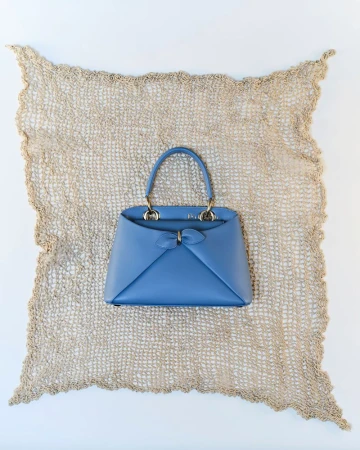 Cornflower blue leather top handle handbag displayed on a beige crochet mat against a clean white background.