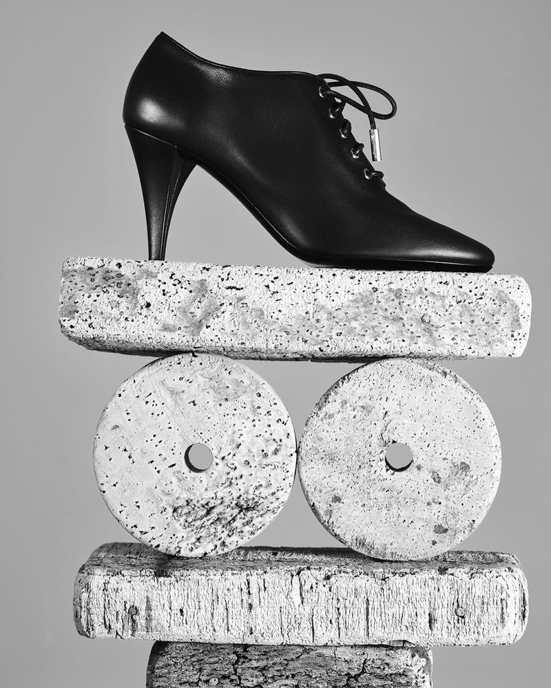 Black leather lace-up heeled shoe balanced on a sculptural stack of textured concrete blocks and discs in studio.