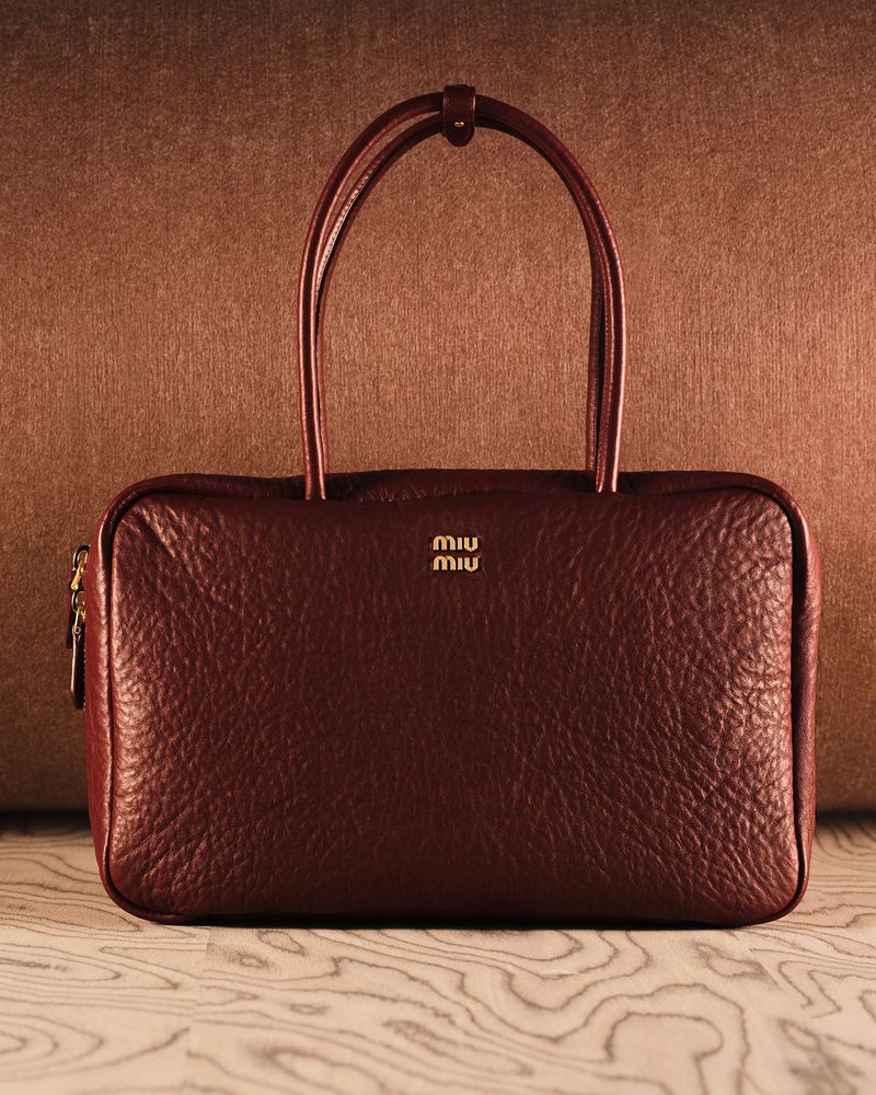 Maroon leather Miu Miu handbag with dual top handles and gold logo, resting on a light woodgrain surface against a textured brown backdrop.