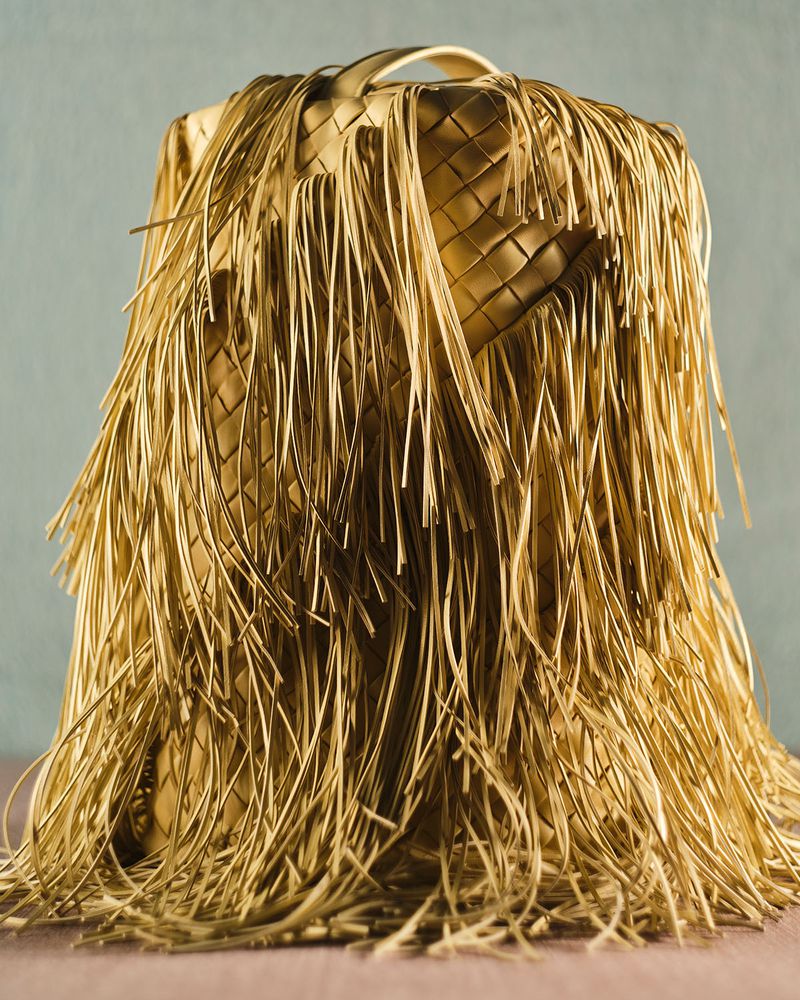 Metallic gold woven leather handbag covered in long cascading fringe on a flat surface against a textured backdrop.