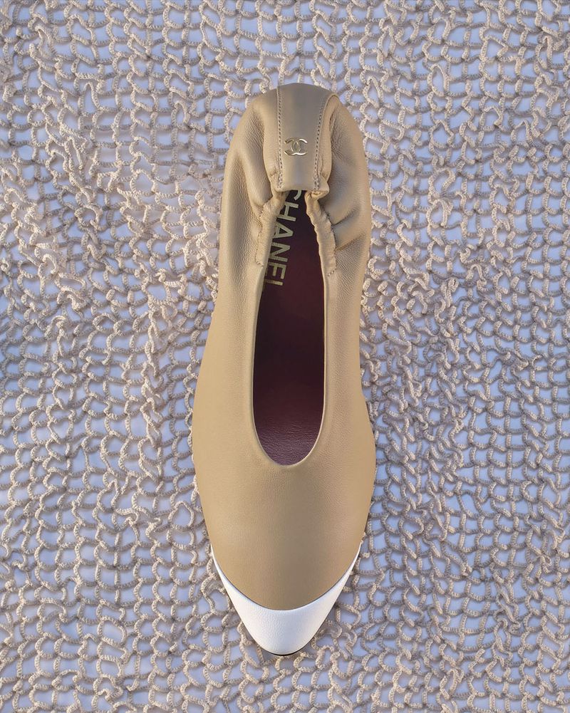 Overhead view of a beige leather Chanel ballet flat with a white cap toe resting on an open-knit mesh fabric background.