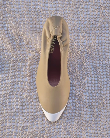 Top-down view of a beige Chanel leather ballet flat with a white toe cap resting on a loose knit mesh textile.
