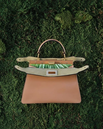 Tan leather handbag resting open on a textured bed of green moss in a top-down view.