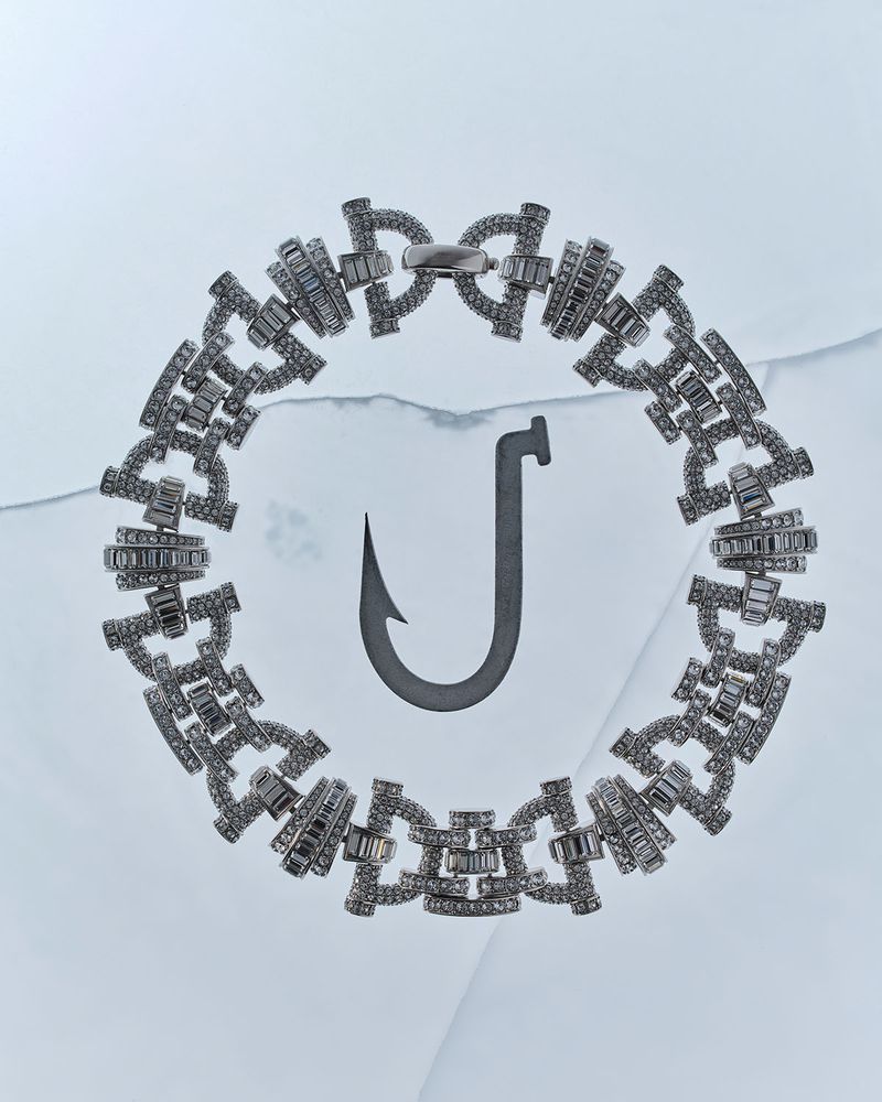 A sparkling diamond chain bracelet encircles a dark metallic fishing hook resting on a pale blue cracked ice surface.