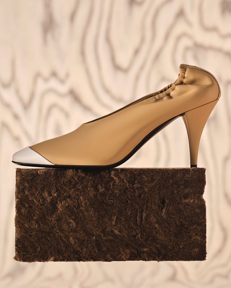 Tan leather high heel shoe with a silver pointed toe cap resting on a dark fibrous block against a wood grain background.