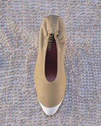 Overhead view of a beige leather Chanel ballet flat with a white cap toe resting on an open-knit mesh fabric background.
