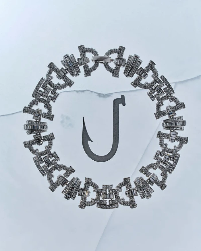 A sparkling diamond chain bracelet encircles a dark metallic fishing hook resting on a pale blue cracked ice surface.