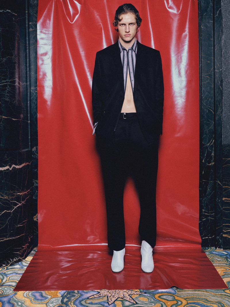 Model Siegfried Blanche wearing an open black suit and white boots standing before a glossy red vinyl backdrop.