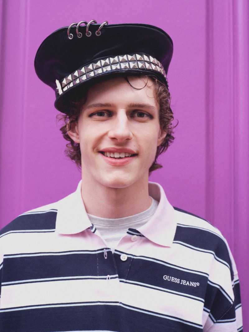 Model Siegfried Blanche smiling in a studded peaked cap and striped Guess Jeans polo shirt against a purple background.