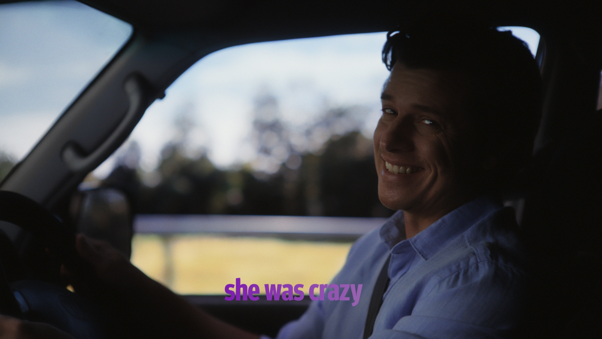 Smiling adult male driving a car in daylight with a rural field in the background, wearing a light blue shirt.
