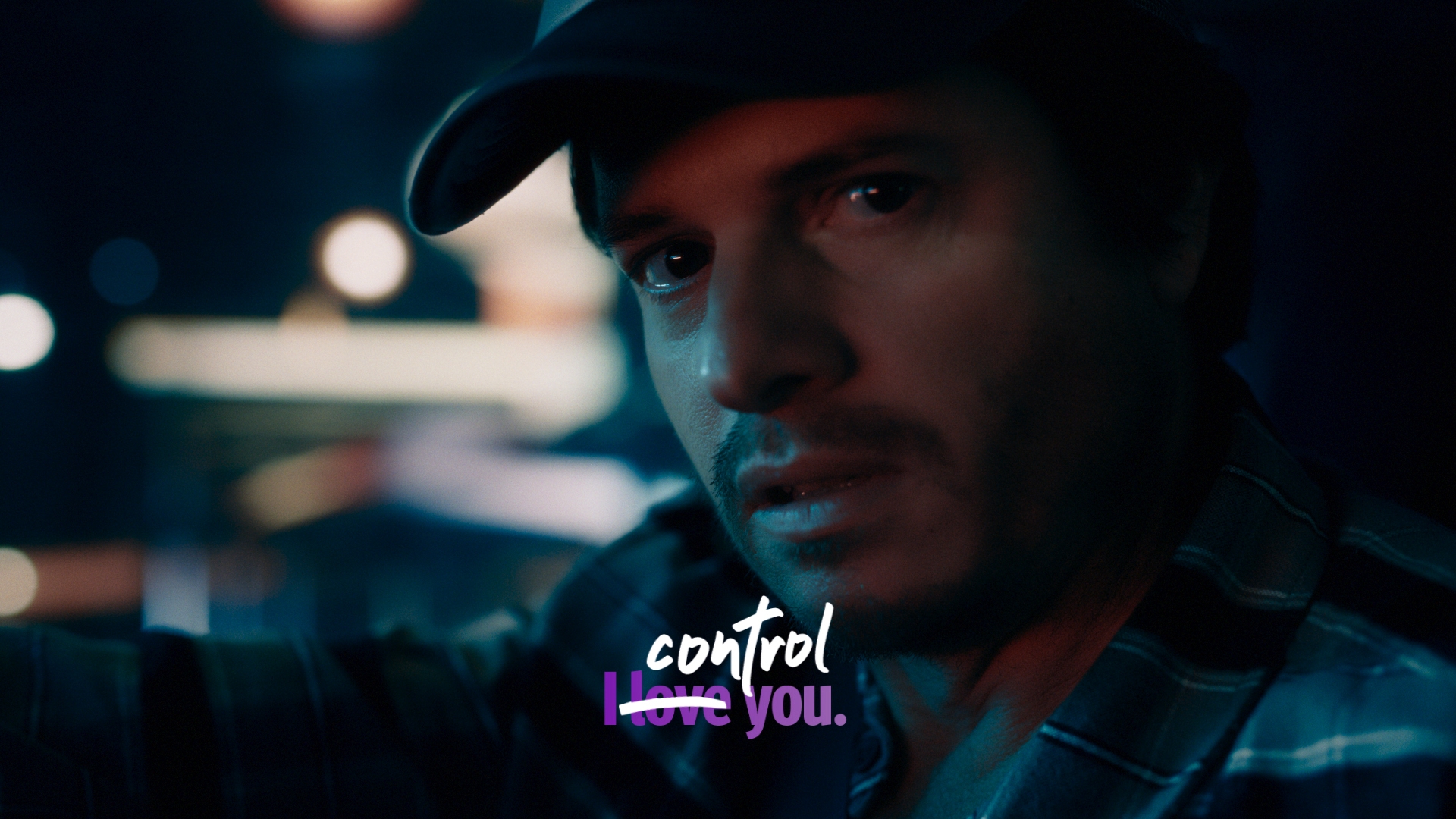 Close-up of an adult male wearing a cap and patterned shirt, looking at the camera in a dimly lit, atmospheric environment.