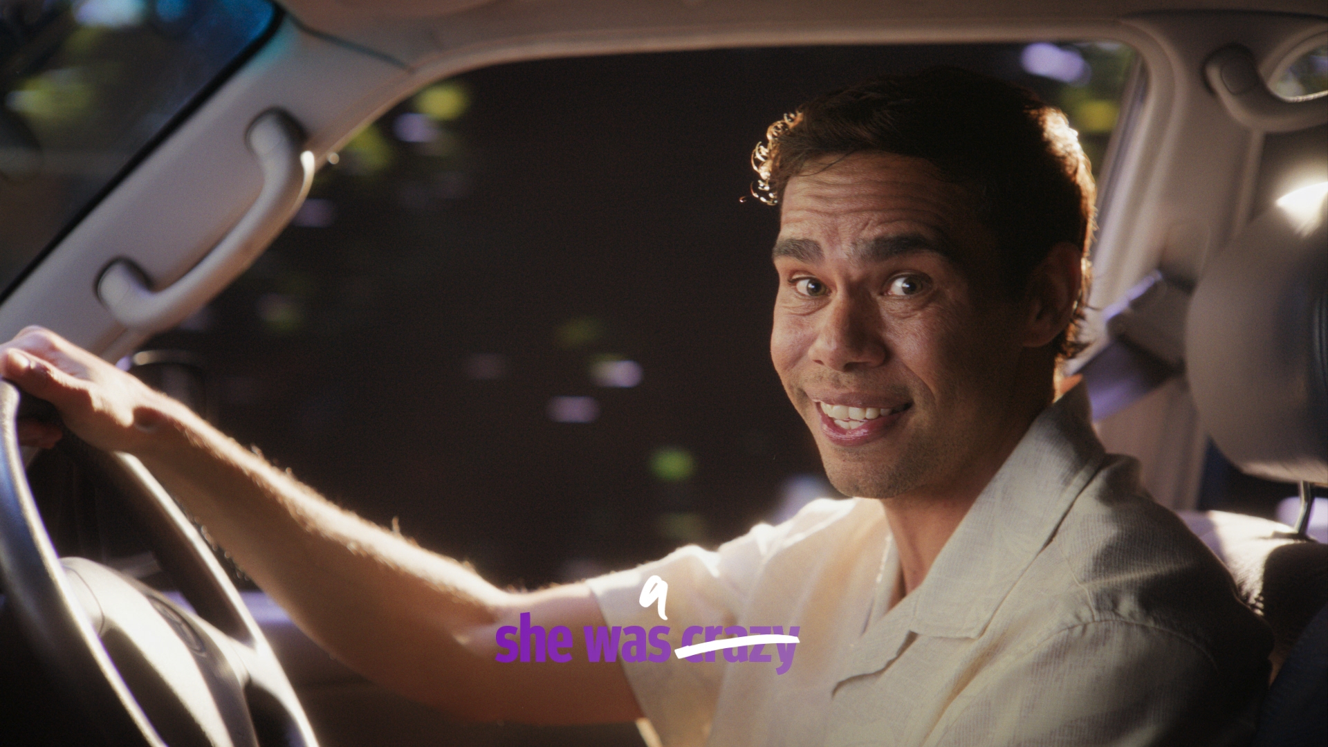Man driving a car, smiling with one hand on the steering wheel, wearing a light-colored shirt, with greenery visible in the background.