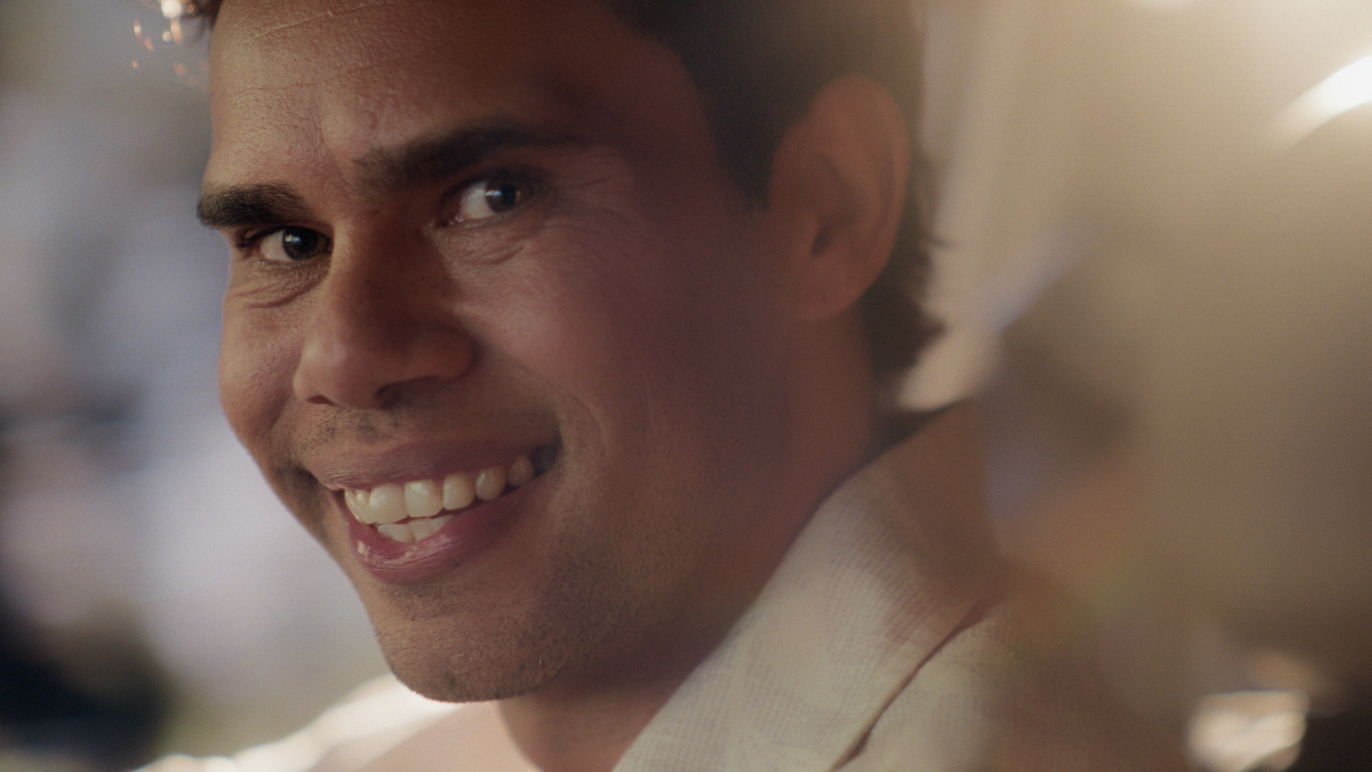 Close-up of an adult male smiling with a white shirt and short dark hair, set against an out-of-focus background with soft lighting.