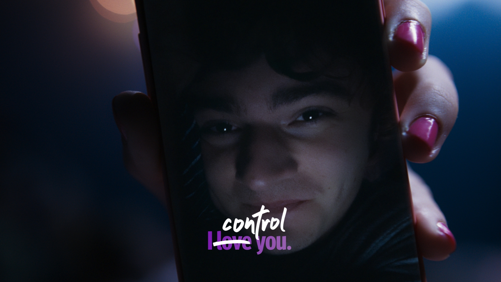 Close-up of a hand holding a smartphone displaying a young man's face with the text 'control' and a crossed-out 'I love you'.
