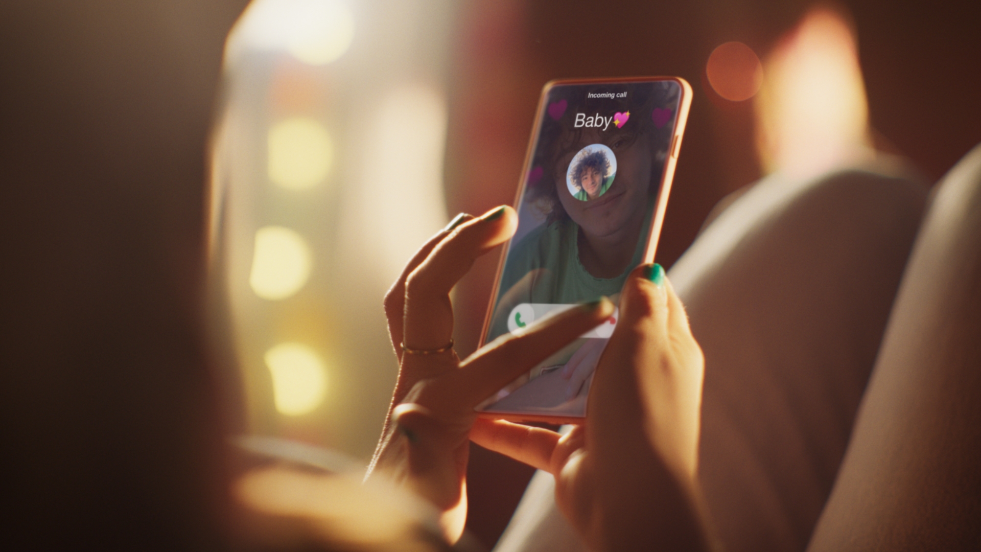 Woman holds smartphone displaying an incoming call from 'Baby' with a heart emoji, illuminated by soft, warm lighting.