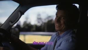 A smiling man driving a car with a light blue shirt, looking back toward the passenger seat.