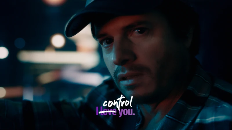 Man wearing a baseball cap and plaid shirt looking intensely toward the camera under dim moody lighting
