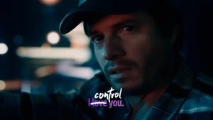 Man wearing a baseball cap and plaid shirt looking intensely toward the camera under dim moody lighting