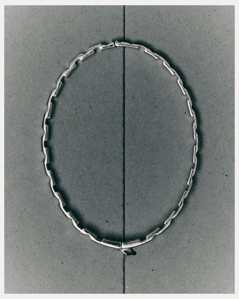 Close-up of a modern, silver-colored chain necklace with rectangular links, displayed on a gray surface with a vertical line running through the center.