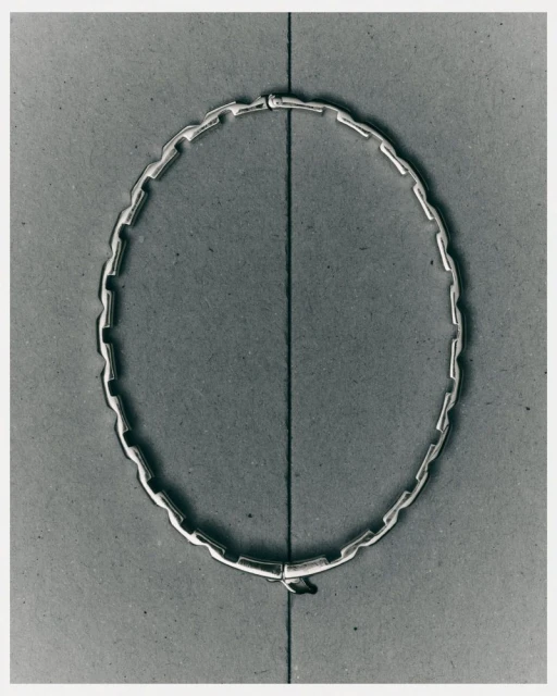 Close-up of a modern, silver-colored chain necklace with rectangular links, displayed on a gray surface with a vertical line running through the center.