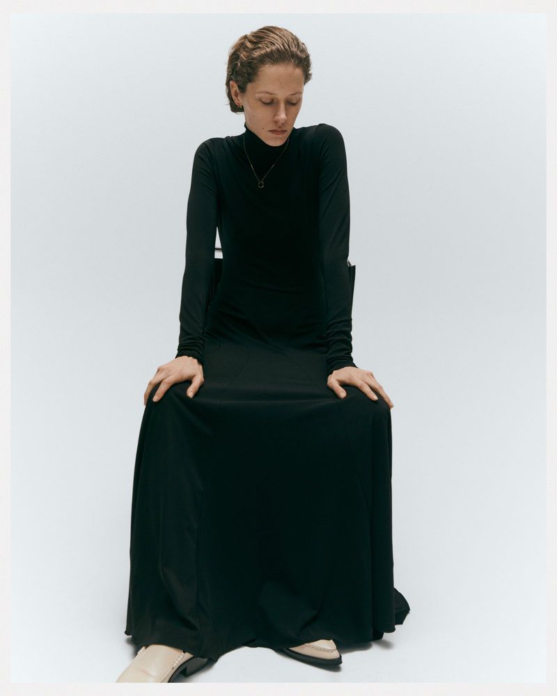 Adult woman in a long-sleeved black dress sitting on a chair in a studio. She is looking down with a neutral expression.