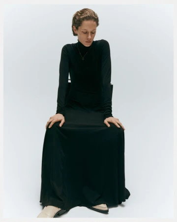Adult woman in a long-sleeved black dress sitting on a chair in a studio. She is looking down with a neutral expression.