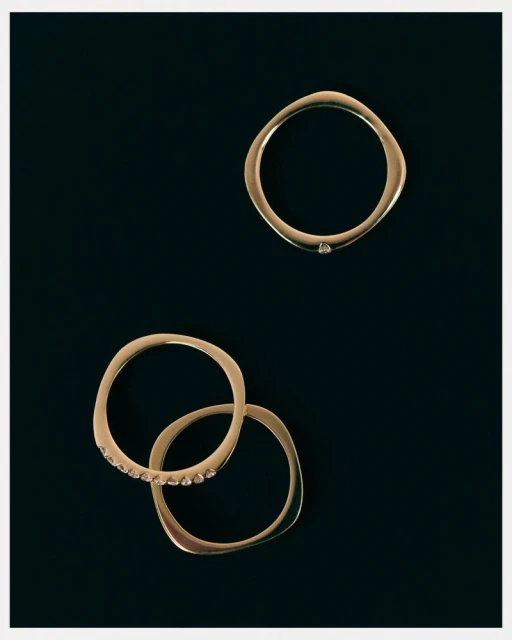 Three gold, rounded rings are displayed on a dark background, one featuring small diamonds, arranged in an aesthetic composition.