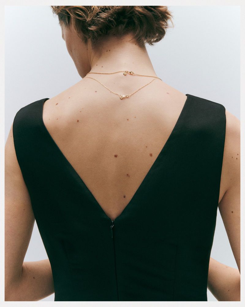 Back view of an adult female wearing a black sleeveless dress and gold necklaces against a white background in a studio setting.