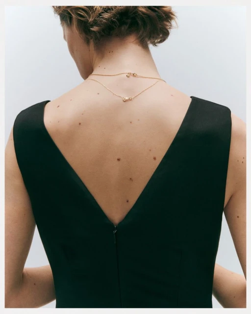 Back view of an adult female wearing a black sleeveless dress and gold necklaces against a white background in a studio setting.