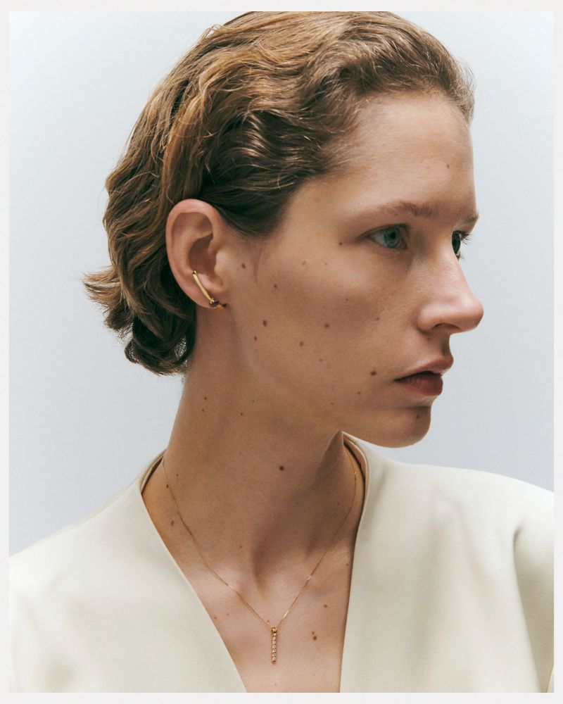 Close-up studio shot of a young adult female with short wavy hair wearing gold jewelry, a necklace, and an ear cuff on a white background.