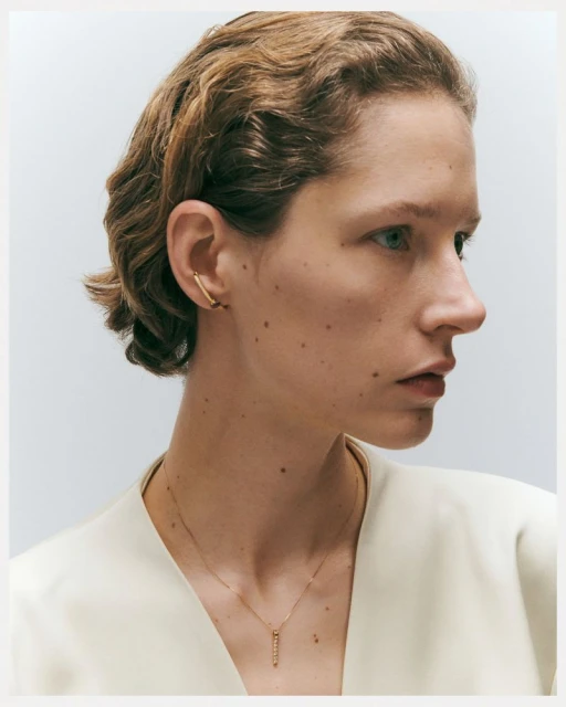 Close-up studio shot of a young adult female with short wavy hair wearing gold jewelry, a necklace, and an ear cuff on a white background.