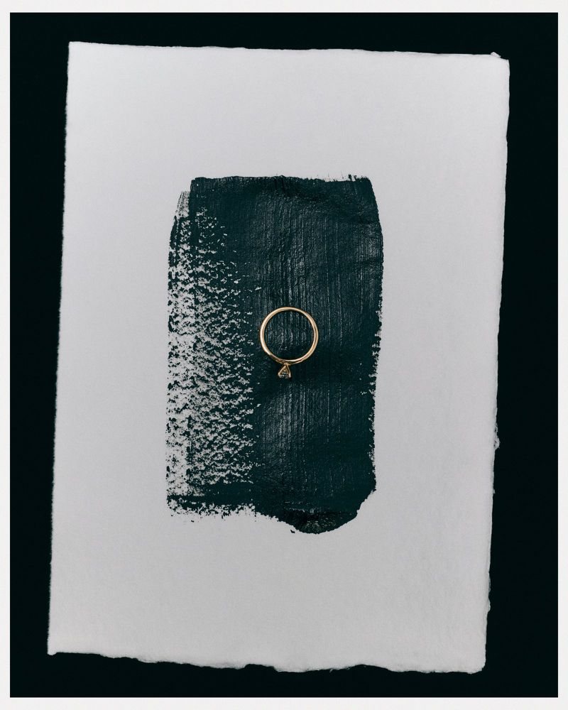 Elegant gold ring displayed on a textured white paper background with a stroke of dark green paint. Minimalist jewellery shot.