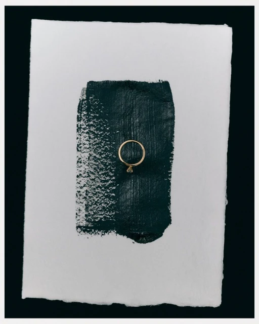 Elegant gold ring displayed on a textured white paper background with a stroke of dark green paint. Minimalist jewellery shot.