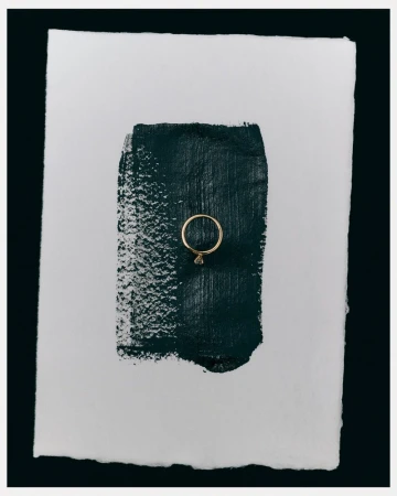 Elegant gold ring displayed on a textured white paper background with a stroke of dark green paint. Minimalist jewellery shot.