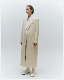 Model wearing a long cream coat and light boots against a plain white studio background.